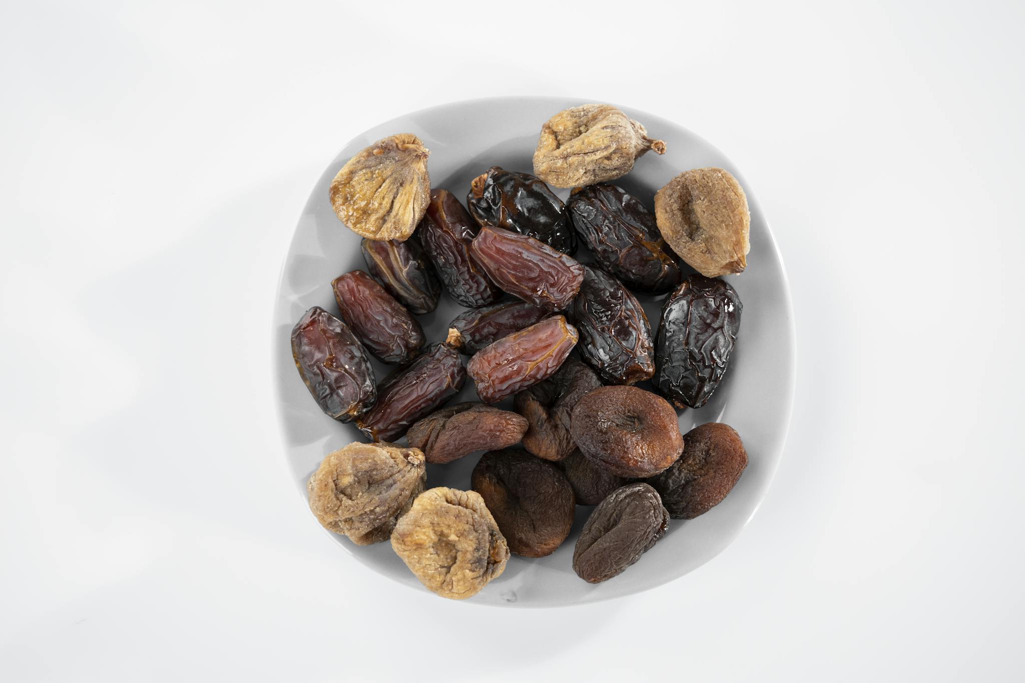 A white plate filled with a variety of dried fruits including dates, figs, and apricots, isolated on a white background.